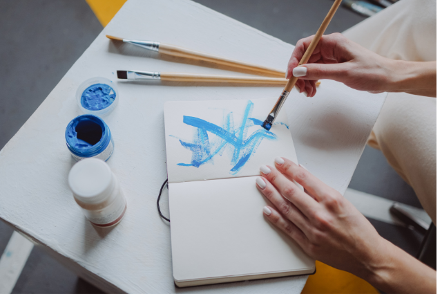 Hands painting abstract blue brushstrokes in a journal with open paint jars on a white table.