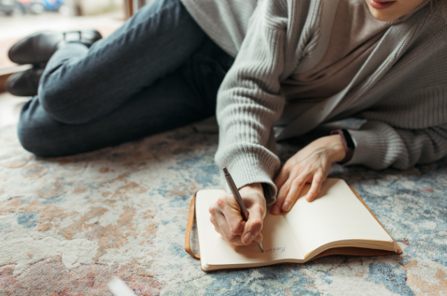 Woman journaling on a patterned rug, wearing a gray sweater and writing with intention in a blank notebook.