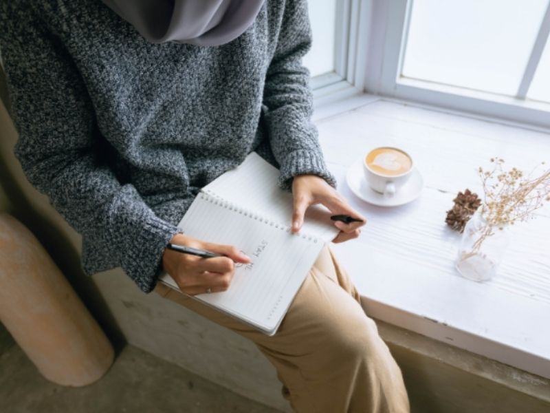 Woman journaling beside a window with coffee and sunlight, practicing journaling for mental health and clarity