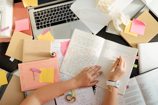Woman journaling at a cluttered creative desk with open notebooks and art supplies, promoting art journaling blog content. Overhead shot organizing notes and study materials on devotionals and art materials. Creative workspace with journals and notes from an art journaling blog, linked from the art journaling disclaimer page.