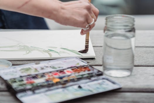 Art print business terms and conditions represented by a hand painting with watercolor beside a palette and jar of water. Artist's hand painting with watercolors in natural light. Creative process captured outdoors.